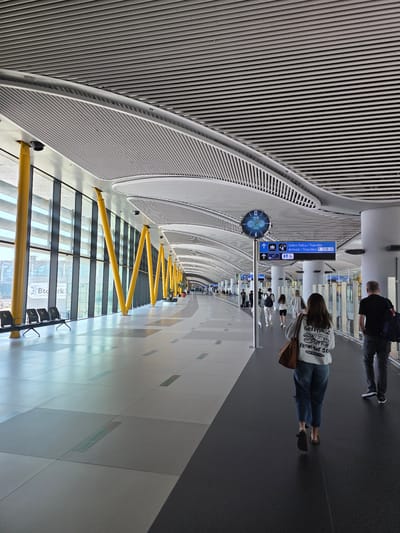 The image depicts a brightly lit interior space, likely within the Istanbul Airport in Arnavutköy, Turkey. A long, wide corridor extends into the distance, with a distinctive curved, ribbed ceiling dominating the upper portion of the frame. A clock and a sign with transportation icons hang beneath the ceiling. Passengers are visible walking along the corridor. On the left side of the image, a row of yellow, zig-zag-shaped supports lines a window. Seating can be seen near the window. The floor is composed of alternating dark and light colored tiles.