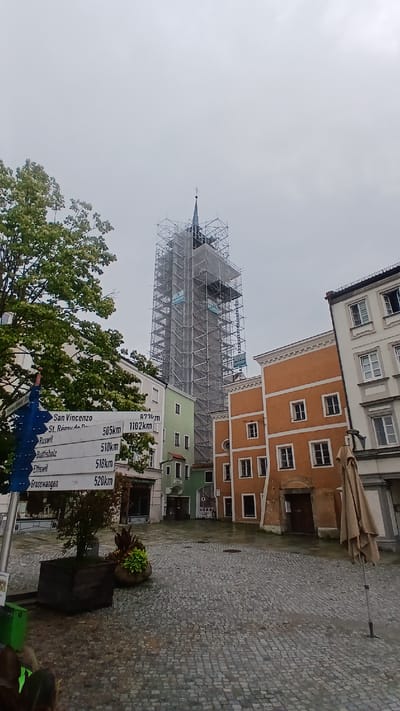 This is a portrait-oriented, full shot of a European town square in Pfarrkirchen, Germany, on an overcast day. The primary focus is a tall building undergoing extensive scaffolding work, likely a church or historic tower, with a spire visible at its peak. Surrounding it are several buildings with colorful facades – one orange and two green – featuring traditional European architecture with multiple windows. The ground is paved with cobblestones, and a large, closed patio umbrella stands near one of the buildings, suggesting an outdoor seating area for a cafe or restaurant.
In the foreground, a large, leafy tree partially obscures the view, and to its right, a series of blue directional signs are visible. These signs list several town names and their distances, indicating this is a public thoroughfare. The text on the signs is partially obscured but includes "San Vincenzo," "St. Rémy de P.," "Ruswil," "Buttisholz," "Ettiswil," and "Grosswangen," along with corresponding distances in kilometers.
The overall atmosphere is subdued due to the overcast weather. The scaffolding suggests ongoing construction or renovation, which is the main activity occurring in terms of the scene's visual narrative. No people are clearly visible in the image. The time of day appears to be daytime, but the lack of direct sunlight makes it difficult to pinpoint precisely. The location is identifiable by the town square setting and the German town name.