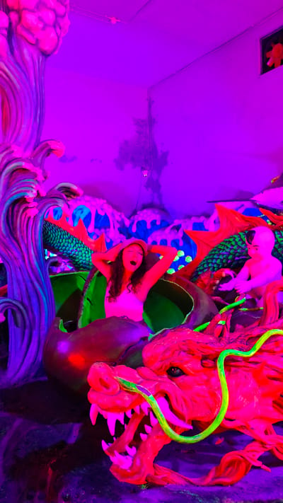A girl is captured in a brightly lit, neon-colored indoor scene, likely at a tourist attraction in Binh Lu, Vietnam. She stands inside what appears to be a dragon's shell, her hands behind her head. The environment is filled with faux waves, and a large red dragon statue dominates the lower part of the frame. A smaller statue of a figure sits behind the dragon. The setting suggests a whimsical or fantastical display. The illumination is primarily purple and pink, creating an artificial and highly saturated atmosphere.