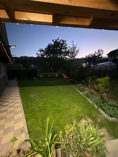 This image captures a serene residential garden or backyard scene at dusk or early evening, set in Kornitsa, Bulgaria. The sky transitions from a soft light blue at the top to a deeper twilight blue towards the horizon, indicating the fading daylight.

In the left foreground, a warm-toned wooden structure, possibly a pergola or part of a house exterior, frames the view. Small, bright string lights are visible along its underside and continue along a roofline further back. A large tree, mostly silhouetted against the sky, stands prominently in the mid-ground on the left.

The central part of the image is dominated by a well-maintained grassy lawn, illuminated by unseen light sources, giving it a vibrant green hue. Along the right side of the lawn, a paved pathway or patio features a distinct checkered pattern of light and dark squares or rectangles. Potted plants with lush green foliage are visible in the immediate right foreground.

Beyond the lawn, along its far edge, is a garden bed containing various smaller plants and shrubs, with several decorative lights glowing softly, casting warm accents. In the deeper background, distant buildings or structures are dimly visible or appear as dark silhouettes against the evening sky. No specific activity is taking place, contributing to the tranquil and peaceful atmosphere of the scene. There is no visible text in the image.