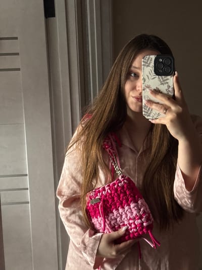 A female subject is depicted in an indoor setting, identified as Tlustenkhabl, Russia. She has long, dark brown hair and is wearing light pink, button-up sleepwear. Her right hand holds a dark-colored smartphone, equipped with a cream-colored, floral-patterned case, positioned to capture a mirror selfie, obscuring the right side of her face. A ring is visible on her left ring finger. Her left hand holds a knitted bag featuring horizontal stripes of magenta and light pink yarn. The bag is square-shaped, has a metal chain strap intertwined with pink material, and a small metallic tag on its front. The background consists of a light-colored, paneled door or wall with vertical grooves.