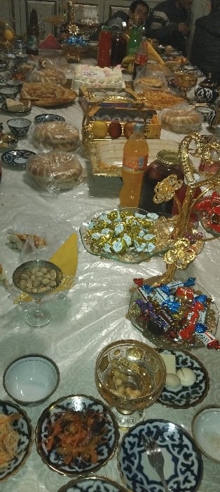 This image depicts an indoor gathering in Fayziabad, Uzbekistan, centered around a long table laden with an extensive variety of food and drink. The table is covered with a white, crinkled plastic tablecloth.
In the foreground and midground, numerous dishes and beverages are arranged. Visible items include several round flatbreads, some individually wrapped in clear plastic. A white frosted cake with pink and white decorations is centrally placed. Assortments of sweets are prominent, including a glass dish filled with chocolates in gold wrappers and a multi-tiered gold-colored stand holding various individually wrapped candies. Another stand holds what appear to be three pomegranates and two lemons. Small bowls and decorative plates, many featuring blue and white traditional Uzbek patterns, hold nuts (pistachios in a silver-colored goblet, other nuts in a gold-patterned glass goblet), a shredded orange vegetable dish, and small white pastries or cookies. Bottles of soda or juice in orange, red, and green colors are interspersed among the food. A dark liquid in a decorative glass jar is also present. A small fork rests on one of the patterned plates.
In the blurred background, partially visible figures of at least two individuals, presumably guests, are seated or standing behind the table. The overall scene suggests a celebratory feast or meal.