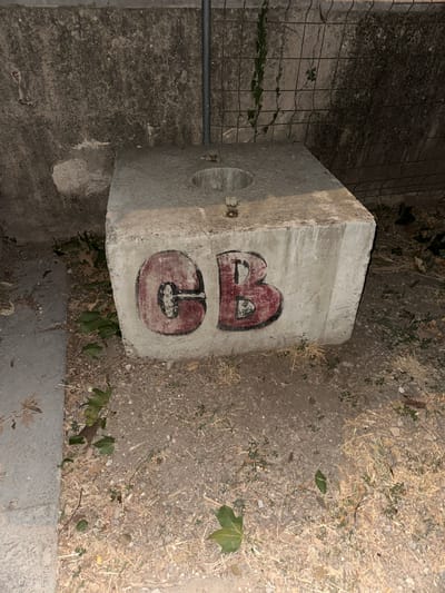A concrete cube with a circular opening on top sits on the ground. The letters "CB" are spray-painted in red and black on the side of the cube. The cube is situated outdoors in a dry, dusty environment with scattered leaves and dry grass. A concrete wall and a metal fence are visible in the background. The lighting suggests it is either nighttime or a dimly lit area. The location is identified as Anduze, France.
