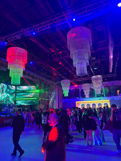 The image depicts a vibrant indoor event space illuminated by a mix of colorful stage lighting, predominantly blue and red, with prominent chandeliers hanging from the ceiling. The environment appears to be a large hall, possibly a venue for a party or concert, with a stage set up on the left side featuring DJ equipment and musical instruments, and large screens displaying abstract, futuristic graphics. A crowd of people are seen mingling and dancing on a polished floor. The lighting creates a dynamic and energetic atmosphere. The scene is set within the city of Akhalubani, Georgia, though no specific geographical landmarks are visible to confirm this location.