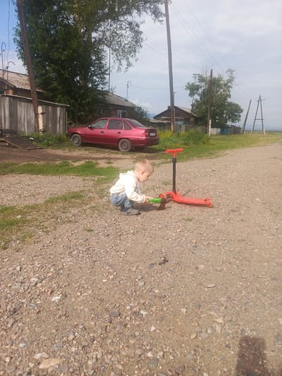In the village of Mezhegey, Russia, a young boy wearing a light-colored hooded sweatshirt and blue jeans squats on a gravelly dirt path. He is playing with a red scooter, and has an axe in his hands. The scooter is red with a black pole and handle. In the background, a red car is parked in front of a wooden fence and a rustic-looking house with a tin roof. Trees and utility poles are visible, along with a cloudy sky.