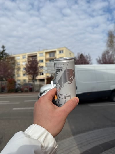 A close-up, first-person perspective photograph captures a right hand holding a silver Red Bull energy drink can and a lit cigarette. The hand, exhibiting light skin tone, holds the can with the thumb and fingers, while the lit cigarette is positioned between the index and middle fingers. The can displays the Red Bull logo (two bulls) and French text indicating "Édition," nutritional information (191 kJ / 45 kcal per 100 ml), and mentions "cerise" (cherry) as a flavor component, suggesting a specific edition. The wrist is covered by a white fabric cuff, possibly from a jacket or shirt.

The background is out of focus, depicting an urban street scene in Lingolsheim, France, under a partly cloudy sky with scattered white clouds. A prominent multi-story residential building, light yellow or beige in color, with multiple windows and balconies, stands in the mid-ground. Deciduous trees, largely bare, are scattered between the viewer and the building, alongside some reddish shrubbery. A paved road surface with faint white lane markings is visible in the lower left. Vehicles include a white panel van partially visible on the right side of the frame, and a silver-grey passenger car discernable on the left side of the road, further down the street. An additional darker, blurred vehicle is present in the distant background.
