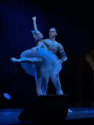 A male ballet dancer wearing a patterned white and grey top holds a female dancer in a dramatic pose. The woman, in a white tutu and tiara, arches backward, her arm extended. A blue light illuminates the scene. Parts of a speaker and what appears to be a stage background are visible on the right. The setting is a dark stage, likely a performance.