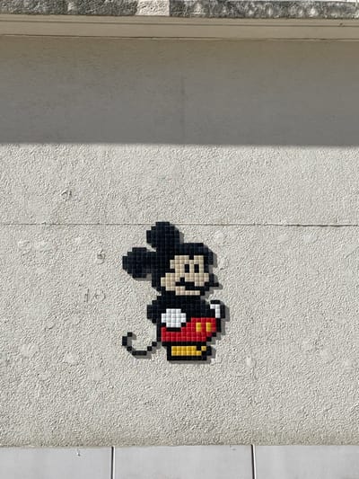 The scene depicts a public art installation on an exterior building wall in Villeurbanne, France. A pixel art mosaic portraying Mickey Mouse, composed of black, beige, white, red, and yellow square tiles, is affixed to a light-colored, granular stucco surface. The mosaic, approximately 30-40 tiles in height, is positioned left of center, casting a sharp shadow to its right and below due to direct illumination from the left. The textured wall is bordered above by a smoother horizontal band leading to a dark grey, textured overhang, and below by a smooth, light-toned base composed of two rectangular panels. No people or dynamic elements are present.