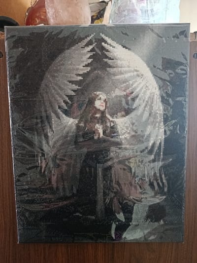 The image shows a framed diamond painting of an angel with large white wings spread wide. The angel has brown hair and is looking down with red lips. The background is dark and appears to be a forest. The diamond painting is wrapped in plastic and is propped against a wooden surface. There are some blurry objects in the background above the painting, including what appears to be a large pink crystal. The scene is indoors, and the overall impression is of a finished craft project. The image was taken in Төмөнкү Ала-Арча, Kyrgyzstan.