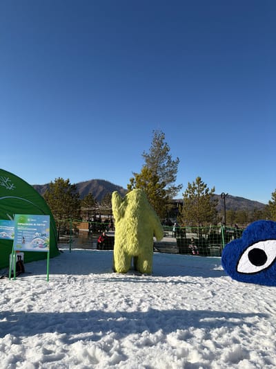 The scene depicts a snowy outdoor area in Озёрное, Russia under a clear blue sky. There are two large, furry character figures present - one blue with a prominent eye and another yellow. A green tent partially obscures the lower left, with a sign in Russian attached. Behind the figures and signage is a metal fence, trees, and blurred figures that appear to be people. A mountain can be seen in the background. The ground is covered in snow.