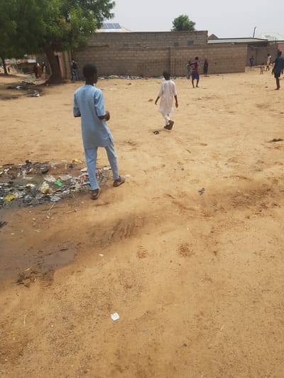 The image depicts an outdoor scene in what appears to be the city of Dutsinma B, Nigeria. The ground is primarily sandy with patches of dirt. A significant pile of garbage and stagnant water is visible in the lower left foreground.
Several individuals are present. A man in a light blue outfit stands near the garbage. Further in the background, another individual is wearing white, and what looks like children are playing a game in sportswear. A brick wall with a tree behind it is also visible in the background.