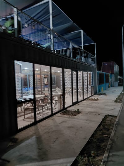 This is a nighttime, eye-level, medium shot of a modern cafe or restaurant built into shipping containers in Akshi, Kazakhstan. The building has a dark, rectangular structure with large glass windows that reveal an interior illuminated by artificial lights. Inside, tables and chairs are visible, suggesting a dining area. Above the main structure, a second level with a railing and a blue canopy is present. To the right of the building, a paved walkway runs alongside a strip of dark earth with some sparse vegetation. Further to the right, other structures, including a dark red shipping container and a brightly painted blue and yellow wall, are visible in the background, along with a street light. The scene is dimly lit, with shadows cast across the pavement.