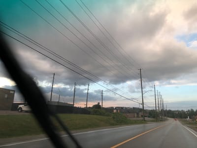 A cloudy sky with a turquoise hue is visible above a rural industrial area. Power lines stretch across the frame, supported by utility poles. A white pickup truck is parked in front of a brick building with the number "500" above its entrance. The scene appears to be on the outskirts of Barrie, Canada, with a wet asphalt road in the foreground, indicating recent rain. The image was likely taken from a vehicle.