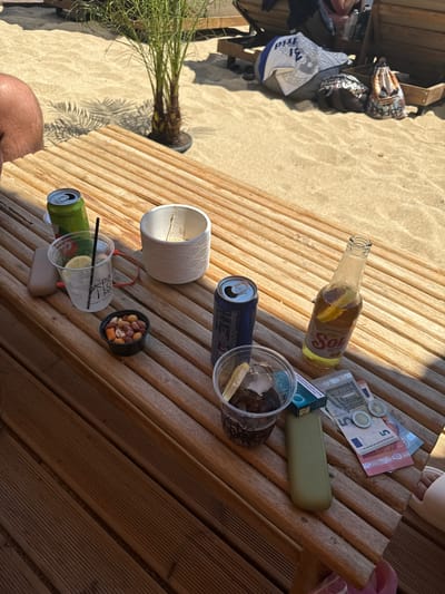 A close-up, eye-level shot captures a wooden table on a sandy beach. The table is populated with several items, suggesting a relaxed, seaside setting. In the foreground, a clear plastic cup filled with ice and a dark beverage, possibly cola, sits next to a blue cigarette pack. To the right of the cup, a bottle of Sol beer, garnished with a lemon slice, leans against the table. Nearby, two Euro coins and several Euro banknotes are scattered, hinting at a recent transaction or simply being left on the table. A green phone case rests beside the cigarette pack. To the left, a green can of a beverage, possibly a soda, is open, and a clear plastic cup with a lemon slice and straw contains a light-colored drink. A small black bowl of mixed nuts is also present. In the background, a potted palm tree stands tall, casting shadows on the sand, and portions of beach loungers and other beachgoers are visible. The overall impression is one of leisure and enjoyment on a sunny day at the beach. The location is identified as Unknown, Greece.