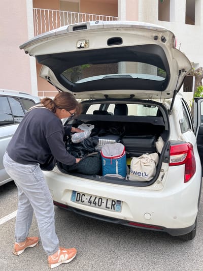A woman with brown hair in a ponytail is packing luggage into the open trunk of a white Citroen passenger car. The car's license plate is DC-440-LR and indicates the car's point of sale: the city of Saint-Etienne. Several suitcases, a canvas bag, and a blue backpack fill the rear trunk. The woman wears a dark sweater, light-colored jeans, and pink and white sneakers. She stands on an asphalt surface, with a building visible in the background. The setting is the commune of Grimaud, France.