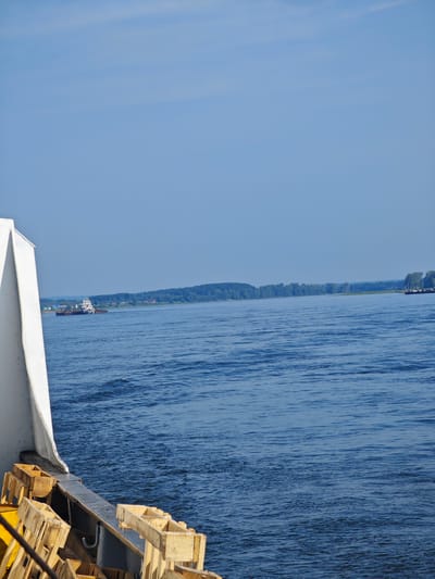 This image depicts a wide, calm river under a clear, pale blue sky. The perspective is from the deck of a vessel, with the white hull of the boat and some wooden crates occupying the foreground on the left side. In the distance, across the river, a green, tree-covered shoreline is visible. A settlement of houses with red roofs can be seen nestled among the trees on the far bank. A tugboat is towing barges on the river, moving towards the left of the frame. The water is a deep blue with gentle ripples. The location is identified as Piskunovkа, Russia.