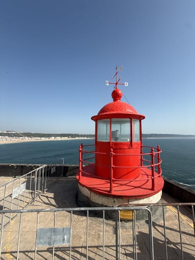 The image depicts a bright red lighthouse, prominently positioned on a cliff overlooking a body of water, likely the Atlantic Ocean. The lighthouse features a bulbous top with a weather vane, and a glass-enclosed section just below. The structure is surrounded by a metallic safety rail, adding a sense of enclosure and protection. The base of the lighthouse is surrounded by a concrete platform.
In the background, a coastal city, presumably Nazaré, Portugal, stretches along the coastline, appearing small in comparison to the dominating presence of the lighthouse in the foreground. The sky is clear and blue. The time of day seems to be midday due to the bright sunlight and shadows, indicating clear weather conditions.