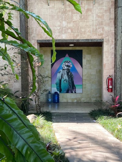The image, seemingly taken from above in Petulu, Indonesia, reveals an interior courtyard space composed of tiled flooring. A painting is displayed on one of the walls, depicting an astronaut figure. Above the painting is a red fire extinguisher. The scene is framed by lush greenery. In the lower right are large green leaves. To the right is concrete and plants. In the lower center are a collection of thin leaved bushes and plants. Two blue water jugs sit on the floor in front of the painting.