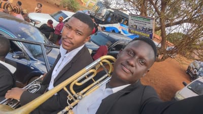 Two men in dark suits pose for a selfie, one holding a trombone. They are standing near several parked cars and a bus on a dirt road. People in traditional clothing are visible in the background, along with a sign hanging from a tree. The overall scene suggests a gathering or event, possibly a celebration. The image is taken outdoors in daylight.