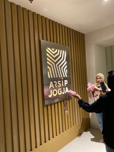 The image shows two women standing in front of a wall with a sign that reads "ARSIP JOGJA" in large, stylized lettering. The sign is illuminated from behind, creating a halo effect. The wall is made of vertical wooden slats. One woman is holding a pink rectangular object in her right hand and is wearing a white hijab and brown top with beige pants. The other woman is wearing a black jacket, dark blue jeans, and a black hijab. She is holding a black smartphone in her left hand. The woman in the black jacket is taking a picture of the other woman holding the pink object. The environment is indoors and well-lit, with beige walls and a door in the background. The location is Banguntapan, Indonesia, at coordinates -7.8 latitude and 110.407 longitude.