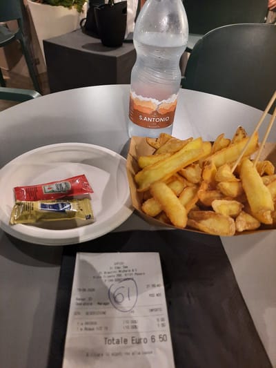 The image shows a close-up view of a table setting with food and drinks, likely in a restaurant or cafe. In the foreground, a white paper plate holds two small packets, one red with "Heinz Tomato Ketchup" written on it, and another yellow packet with some blue writing. To the right of the plate, a cardboard container is filled with french fries. Behind the plate and fries, a bottle of "S. ANTONIO COMO FRIZZANTE" mineral water stands prominently. A receipt is partially visible in the lower center of the image, showing items like "PATATINE" and "Acqua 1/2 lt," with a total of "Euro 6.50." The receipt also indicates the location "PESARO" and a date of "19-06-2020." The overall environment appears to be indoors, with muted lighting and dark furniture in the background.