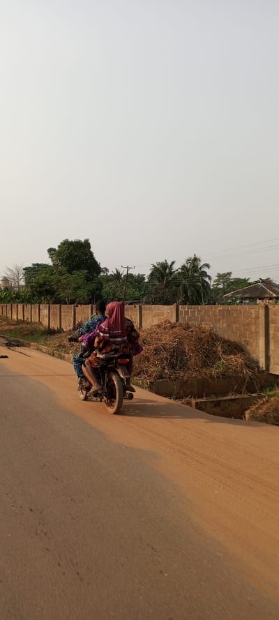 Two individuals are riding a motorcycle on a dusty, reddish-brown road, viewed from the rear. The road appears to be paved in the center but with significant dust and unpaved sections on the sides. Tire tracks are visible on the road surface.

The driver, presumably male, is wearing patterned blue and white clothing. The passenger, seated behind the driver, appears to be female, wearing a red/maroon head covering (possibly a hijab) and similarly patterned clothing in shades of brown, orange, and purple. Both riders' lower legs and feet are visible, suggesting bare feet or sandals. The motorcycle is a dark-colored, utilitarian model.

To the right of the road, a low concrete block wall or fence runs parallel to the road. Behind this wall, there's a large pile of dry straw or brush. Further in the background, a dense line of lush green tropical trees and foliage is visible, along with utility poles and power lines. A faint structure with a roof can be discerned in the far background on the right.

The sky is bright and appears light grey or off-white, suggesting a sunny day, possibly with some atmospheric haze. The overall setting indicates a semi-urban or rural environment, consistent with the provided location context of Ketu Adieowe, Nigeria. There is no visible text in the image.