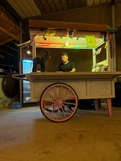 The scene depicts an indoor or covered area, identified as being in the city of Batzra, Israel. The primary subject is a light-colored wooden mobile cart with a counter, equipped with a pink spoked wheel on its left side and a supporting leg on its right. Above the counter, a canopy structure is adorned with a grid of multicolored lights, including red, green, and white/yellow, along with small, triangular decorative flags, some appearing to be prayer flags.

One adult male, approximately 30-40 years old, with short dark hair and a beard, is positioned behind the cart's counter. He is wearing a dark, short-sleeved shirt and is looking downwards toward the counter's surface. On the counter, to the left, stands a dark-colored elephant figurine with its trunk raised. To the right of the man, two metallic, bowl-shaped containers are visible on the counter.

The environment is dimly lit, with artificial lighting originating primarily from the cart's canopy and supplementary hanging lights in the upper left. The floor is a light-colored concrete or paved surface. In the background on the left, a large, dark, oval-shaped object and a blue plastic bag are visible on the ground, alongside a partially visible refrigerator or cooler with internal illumination. On the right, dark crates are stacked near the base of the cart. The ceiling overhead consists of wooden planks and exposed rafters.