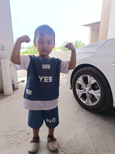 The media file shows a young boy, likely under ten years old, standing outside next to a white car. He is flexing his biceps, with his left arm bent upwards and his fist clenched, and his right arm also bent upwards. The boy is wearing a dark blue t-shirt with a white band around the collar and white trim around the sleeves. The t-shirt has white text that says "YES" printed on it in large letters, with a barcode design above it. He is also wearing dark blue shorts with white text that says "NO" on the left leg. He is wearing light-colored slip-on shoes with brown trim and brown socks.

The scene appears to be an outdoor setting, possibly a driveway or courtyard, with a concrete floor. To the left of the boy, there is a white sink and plumbing fixtures. In the background, there are some green trees and a brick wall. A white car is parked to the right of the boy, with its front wheel clearly visible. The time of day appears to be daytime, with bright sunlight, suggesting fair weather. The boy's expression is neutral. The location is identified as Langar, Uzbekistan.