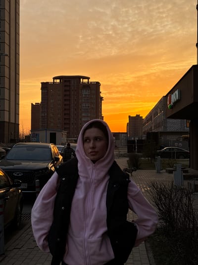 An outdoor urban scene in Gribovo, Russia, captured during sunset, features a young female subject in the foreground. The sky displays vibrant orange and yellow hues with scattered clouds. The female subject, positioned slightly right of center, has light skin and is wearing a pink hooded sweatshirt with the hood up, covered by a dark, possibly black, sleeveless puffer vest. She is looking upwards and to her left, with her hands out of frame.

In the mid-ground, a dark SUV, identifiable as a Chery Tiggo 8 Pro, is parked to the left of the subject, displaying a Russian license plate "550 RUS". Further behind, another male individual in a plaid jacket is visible standing between parked vehicles.

The background is dominated by multi-story residential buildings. A tall, light-colored building frames the left side of the image. Centrally, a prominent dark-red/brown multi-story building with numerous windows and a distinctive top structure stands against the sunset. To the right, a low-rise commercial building, partially under construction, displays a "ДИКСИ" (DIXY) store sign. Sparse, dark bushes and bare trees are present along the paved sidewalk in the mid-ground. A small, light-colored dog is observed walking on the sidewalk in the mid-ground, to the left of the bushes. The ground consists of paved walkways and an asphalt parking area.