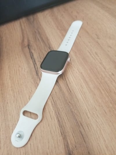 A close-up, high-angle shot shows a white-strapped smartwatch with a dark screen placed on a light brown wooden surface. The watch band is looped, with a circular indentation and a silver stud visible on the lower strap. The wooden surface exhibits a distinct grain pattern with darker lines running diagonally across the frame. The upper left corner of the image is partially obscured by a dark, out-of-focus object. The scene is indoors, with the lighting suggesting an artificial light source. There are no people or animals present. The smartwatch appears to be the sole subject of interest. The location is not identifiable as Usolye-Sibirskoye, Russia, or any specific city.