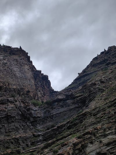 A wide-angle, low-angle shot captures two steep, rocky cliff faces that converge to form a narrow ravine. The sky is overcast with thick, grey clouds. The cliffs are composed of dark, layered rock with patches of green vegetation growing in crevices and on ledges. Two small figures are visible on the upper ridges of the respective cliffs. The overall impression is one of rugged, imposing natural scenery. This scene is located in the vicinity of Gondhla, India, a region characterized by its mountainous terrain.