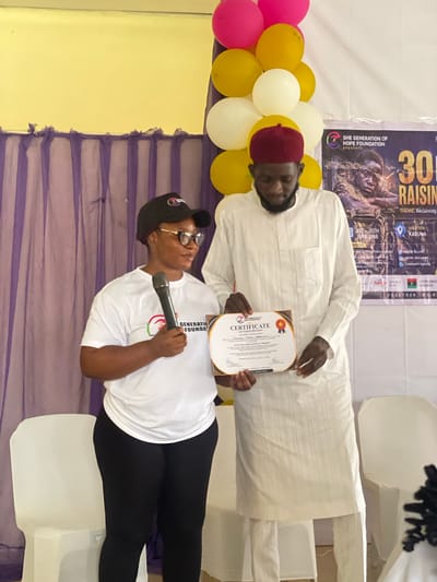 In Unguwar Shanu, Nigeria, a woman in a white t-shirt and black leggings, wearing glasses and a black cap, stands on the left holding a microphone and a certificate. To her right, a man in a white traditional outfit and a red cap is also holding the certificate, which is being presented. Balloons in pink, yellow, and white are visible in the background, along with purple curtains and a banner that reads "SHE GENERATION OF HOPE FOUNDATION PRESENTS 30 R." The scene appears to be an indoor event, possibly a presentation or award ceremony, taking place in a room with white chairs.
