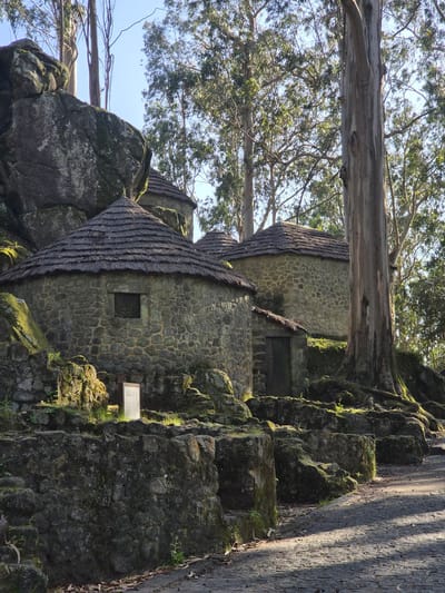 The image shows a stone building with thatched roofs built into a rocky outcrop in Esposende, Portugal. Trees overhang the building. There is a stone path running along the right side of the structure. Some moss grows on the rocks.