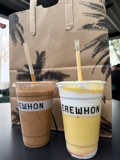 The image shows two Erewhon smoothies on a table with an Erewhon paper shopping bag nearby. The top smoothie is yellow and white, while the bottom smoothie is brown. Each smoothie has a straw. The shopping bag has a palm tree print on it. The location is likely in Pasadena, United States of America, based on the presence of Erewhon.