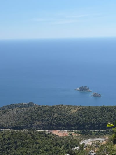 The image displays a scenic vista from a high vantage point overlooking the Adriatic Sea near Novoselje, Montenegro. The foreground is dominated by lush green hills covered in dense vegetation. A winding road snakes its way through the terrain, disappearing into the trees. In the middle ground, the deep blue expanse of the sea stretches out to the horizon. Two small, rocky islands, sparsely covered with trees, rise from the water, adding to the picturesque landscape. The sky above is a clear, bright blue.