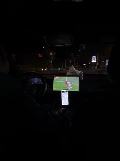 A dark interior shot from a driver's perspective shows a screen displaying a soccer game attached to the dashboard. A smartphone, displaying a navigation app, is mounted next to the screen. The steering wheel and partial dashboard are visible. Reflections of city lights are seen through the windshield. A box of tissues rests near the screen, hinting at other items in the car.