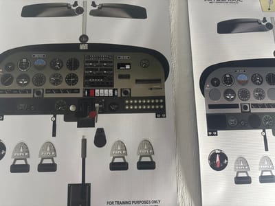Two large posters illustrating aircraft cockpit components are affixed to a light-colored wall in Port Alfred, South Africa. The predominant left poster displays a detailed, full-scale depiction of an aircraft instrument panel, primarily gray with black accents. It features a complete set of flight instruments, including an artificial horizon, altimeter, airspeed indicator, and engine gauges. Communication and navigation radios, toggle switches, and circuit breakers are also present. The registration identifier "ZS-XYZ" is visible twice on the panel. Below the main panel, two silver rudder pedal assemblies, each bearing the text "RUDDER," are depicted alongside a central black control column. A red-handled lever is visible on the lower-right section of the panel. The bottom of this poster contains the text "FOR TRAINING PURPOSES ONLY." A partially visible second poster on the right is similar in style, showing a segment of an aircraft instrument panel with gauges, the identifier "ZS-XYZ," and text at the top indicating "AIR SCHOOL" and "NEXT GENERATION TRAINING."