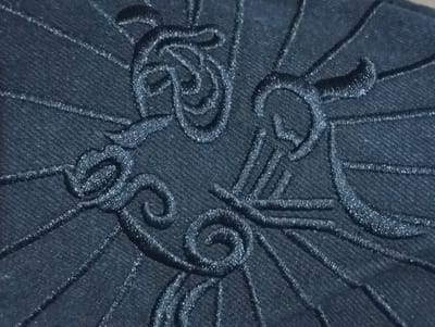 A close-up, high-angle shot shows intricate dark blue embroidery on a dark blue fabric, possibly denim. The embroidery forms abstract, swirling patterns and radiating lines, creating a textured, almost three-dimensional effect against the fabric's weave. The lighting casts subtle shadows, enhancing the depth and detail of the embroidered design. There are no people or discernible environmental context visible, suggesting the image focuses solely on the textile detail. The location in Ukpor, Nigeria, is not depicted or implied by the visual content.