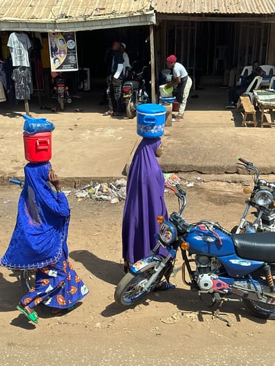 This image depicts a bustling street scene in Tataura, Nigeria, under bright daylight. In the foreground, two women are walking from left to right on a dirt path. The woman on the left wears a blue head covering, a patterned blue and orange dress, and green sandals. She carries a red cooler topped with a blue plastic bag, balanced on her head. The woman on the right wears a purple head covering and a long purple garment. She carries a light blue cooler with a floral pattern, also balanced on her head.
To the right of the women, two motorcycles are parked on the dirt path; the front of a blue motorcycle with a kickstand is prominently visible. The ground between the path and the shops contains scattered litter.
In the background, a row of open-front shops or stalls features a corrugated metal roof supported by wooden poles. On the far left, clothing items, including t-shirts, hang for sale. A poster depicting faces and text is affixed to a wall. Several men are present in and around the shop area. On the left side of the shop, men are standing near parked motorcycles. On the right side, a person wearing a red head covering is bent over, actively washing items in a white bucket. Further to the right, another individual is seated at a wooden table. Strong shadows indicate direct overhead sunlight.