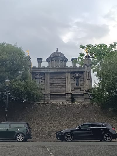 The image depicts a view of the exterior of the Arundel Museum in Arundel, United Kingdom, under an overcast sky. The museum is a stone building with a distinctive dome and ornate architectural details. In the foreground, two cars are parked on the street: a dark green Mini Countryman on the left and a black Mercedes-Benz A-Class on the right. Trees and shrubbery partially obscure the view of the museum. A vintage lamppost stands to the left of the museum. The ground is paved with asphalt and lined with white road markings. The overall scene suggests a quiet street in a historic town.