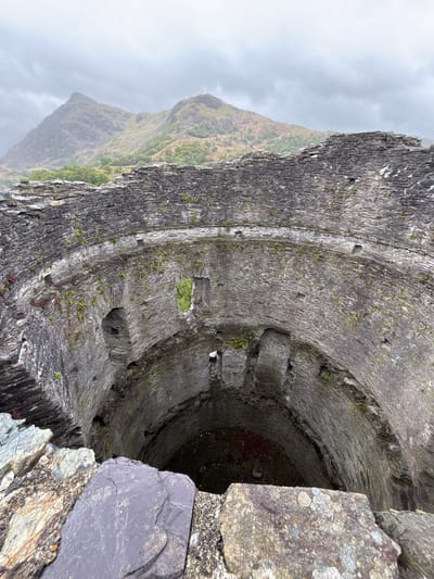 This photograph shows the interior of a slate mine tower in Dinorwic Quarry, Llanberis, United Kingdom. The image focuses on the circular, terraced structure of the tower, built from dark grey slate stones. Moss and small plants grow on the ledges and between the stones. The tower drops into a dark, seemingly bottomless void. To the left, a partially visible mountain fades into the cloudy, overcast sky. The image is taken from a high angle, looking directly down into the tower. The composition emphasizes the depth and texture of the stonework.