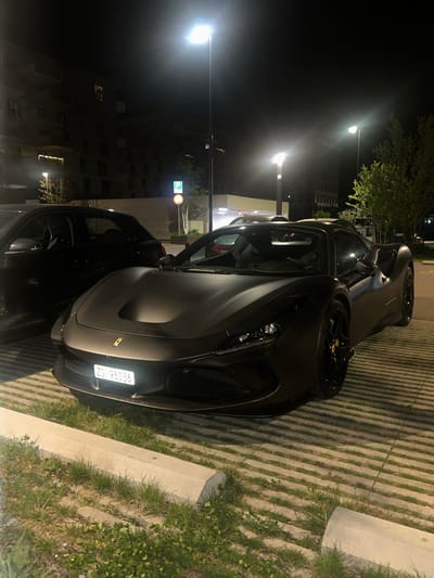 The image captures a nighttime outdoor scene, likely in a parking area in Dübendorf, Switzerland, as suggested by the Swiss license plate format. Two luxury cars are parked side-by-side under the illumination of streetlights.
The primary subject is a sleek, matte dark grey or black Ferrari positioned in the foreground, facing slightly towards the viewer. Its aerodynamic lines, intricate front grille, and modern headlights are visible. The car features large dark alloy wheels with distinctive yellow brake calipers and a small yellow Ferrari emblem on the front hood. A clear white license plate, "ZG 98036", is prominently displayed on the front bumper. "ZG" is the canton code for Zug, Switzerland.
Behind and slightly to the left of the Ferrari, another dark-colored vehicle, possibly a luxury SUV or sports car, is parked. Fewer details are discernible for this second vehicle due to its position and the shadows.
The parking surface consists of permeable pavers with strips of grass growing between the concrete grids, indicating an environmentally conscious design. A concrete curb runs along the edge of the parking area, bordered by more grass and some low-lying foliage. The scene is well-lit by several bright streetlights, which cast a luminous glow and create defined shadows. In the background, multiple-story buildings, possibly residential or commercial, are visible, with some windows illuminated. A blue circular parking sign with a white 'P' is also noticeable in the mid-ground. The night appears clear, with no adverse weather conditions visible.