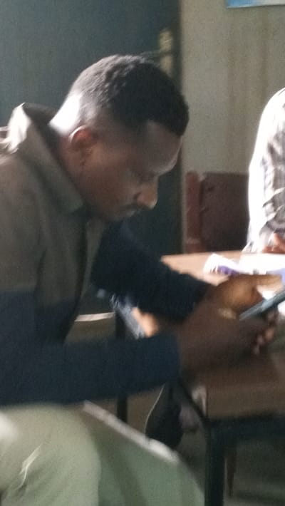 An indoor scene in the city of Dakata, Nigeria, depicts an individual seated at a wooden table, engaged with a smartphone. The primary subject is a male with dark skin and short, dark hair, seen in profile looking down. He wears a light-colored collared shirt over a dark blue long-sleeved shirt. His hands are holding a dark-colored smartphone, which he appears to be operating. On the table in front of him are white and purple papers. A second individual, wearing a plaid or checkered shirt, is partially visible to the right in the background. The environment is dimly lit, with visible furniture including tables and chairs. A dark wall or door is present in the left background, and a lighter wall with a faint, blurry blue-and-white object is visible further right.