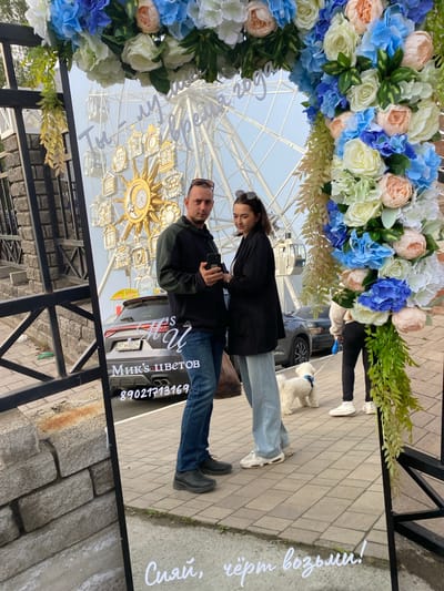 The image, captured in Listvyanka, Russia, shows a couple reflected in a large mirror adorned with a floral arch. The couple, a man and a woman, are standing and looking at a smartphone. The man wears a dark hoodie and jeans, while the woman is in a black jacket and light blue jeans. Behind them, a Ferris wheel is visible, along with cars and a small white dog on a leash being walked by a person in the background. The mirror also displays text in Russian: "Ти - лучшая года" (You are the best year) at the top, and "Мик's ЦВЕТОВ" (Mik's FLOWERS) with a phone number below, indicating a flower shop. Another line of text in cursive reads "Сияй, чёрт возьми!" (Shine, damn it!). The scene takes place outdoors on a paved walkway, with a stone wall and a black metal fence to the left.