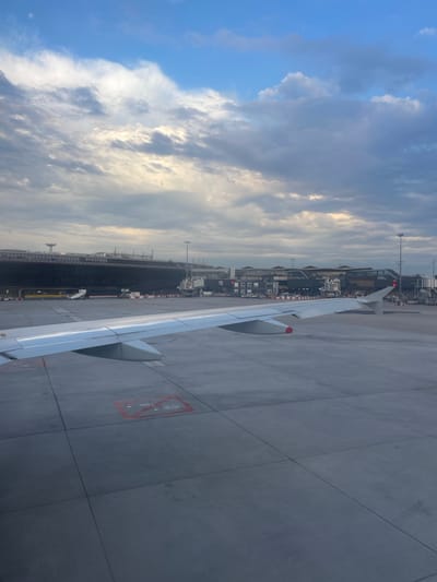 This is an aerial view of an airport tarmac, likely at Paris-Orly Airport in Paray-Vieille-Poste, France. The foreground is dominated by the wing of an airplane, which is silver with red markings on the wingtip. The tarmac is a large, grey, concrete expanse marked with painted lines and symbols, including a red square with two circles and a diagonal line, possibly indicating a no-parking zone or a specific operational area.

In the mid-ground, a large airport terminal building stretches across the frame. It is a multi-story structure with a dark facade and a series of interconnected sections. Jet bridges extend from the terminal to various gates, with several aircraft parked at these gates. Various airport vehicles, such as service trucks and stairs, are visible around the aircraft and terminal.

The background consists of a sky with scattered clouds. Some clouds are dark and ominous, suggesting potentially overcast conditions or the early stages of weather change, while others are lighter and illuminated by sunlight filtering through. The overall impression is a typical scene at a busy airport, likely during the day given the lighting. There are no people clearly visible in the image.