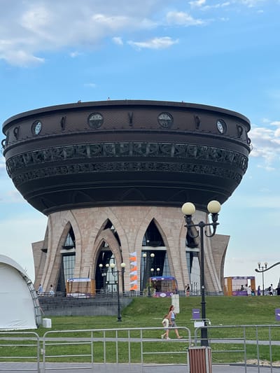 The image shows the "Kazan Family Center," also known as the "Chasha" or "Cauldron," a distinctive building shaped like a giant cauldron, located in Kazan, Russia. The building has a large, dark-colored, bowl-shaped upper section with ornate carvings and circular decorative windows. This rests on a base constructed of light-colored stone with arched openings. In front of the building, there is a grassy area with a fence, a white tent on the left, and lampposts with spherical lights. Several people are visible walking on a pathway in front of the building, some standing near kiosks, and a few are on the roof of the cauldron-shaped structure. The sky is blue with scattered clouds. The specific location of Высокая Гора is incorrect, as this building is in Kazan.