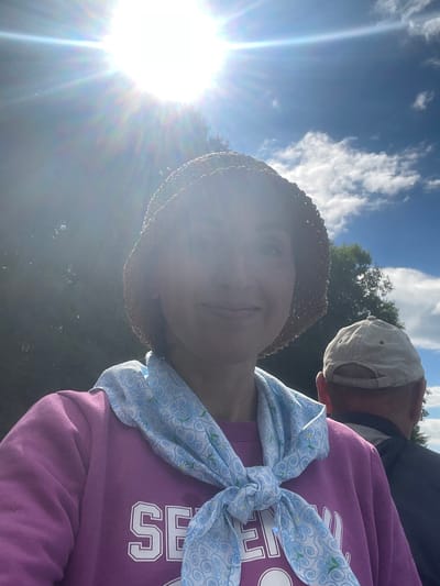 The image is a selfie taken outdoors under a bright sun. A woman wearing a straw hat, a purple sweatshirt, and a blue patterned scarf is in the foreground, looking towards the camera. Behind her, a man wearing a beige baseball cap and a dark jacket is visible from the back. The background shows trees and a blue sky with clouds. There is no information in the image that indicates the location is Dresvyanovo, Russia.