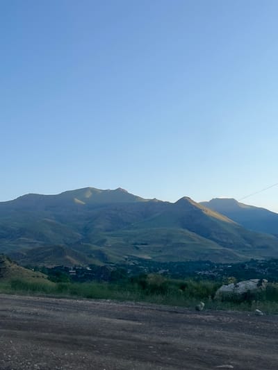 The image depicts a landscape dominated by rolling, green mountains under a clear, pale blue sky. In the foreground, a dirt road or path is visible, with some rocks and sparse vegetation. The middle ground features a densely vegetated slope leading up to the base of the mountains, with several buildings and what appears to be a small village nestled within the valley. The mountains themselves are covered in a mixture of green vegetation and exposed rock, with shadows suggesting the sun is either rising or setting, casting a warm glow on some of the peaks. There are no people or animals visible in the frame. The scene is tranquil and natural, suggesting a rural or mountainous setting, consistent with the location in Arpi, Armenia.
