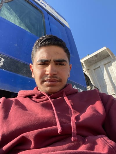 A man with dark hair and a burgundy hoodie stands in front of a blue truck. His face is visible, and he appears to be looking at the camera. The truck's door and a portion of its body are visible, with some writing on the side. Above the truck is a white, rectangular structure. The sky is a clear, bright blue. The angle of the shot is slightly tilted.
