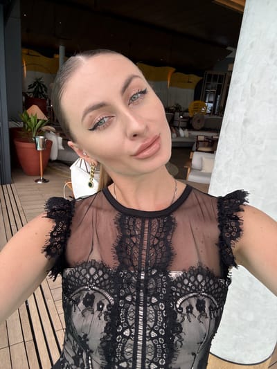 A woman with blonde hair pulled back, wearing a black lace top and gold earrings, takes a selfie. She has on makeup, including winged eyeliner and lip gloss. She is standing on a wooden deck, with a resort-like setting in the background, including potted plants, seating areas, and a white column. The location appears to be outdoors, possibly at a beachside resort in the Maldives, given the tropical vegetation and open-air architecture.